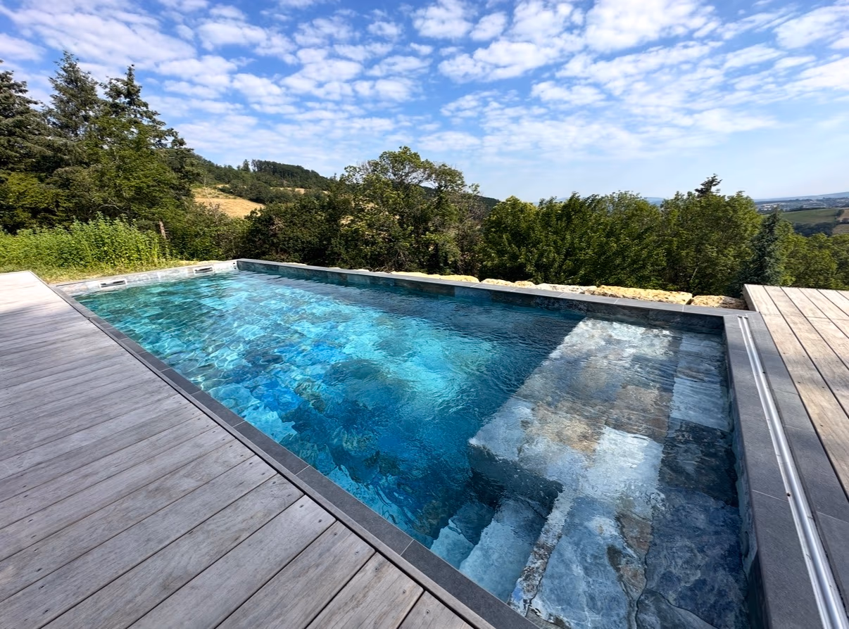 Piscine extérieure habillée de carreaux de pierre bleue et grise, bordée d’une terrasse en bois