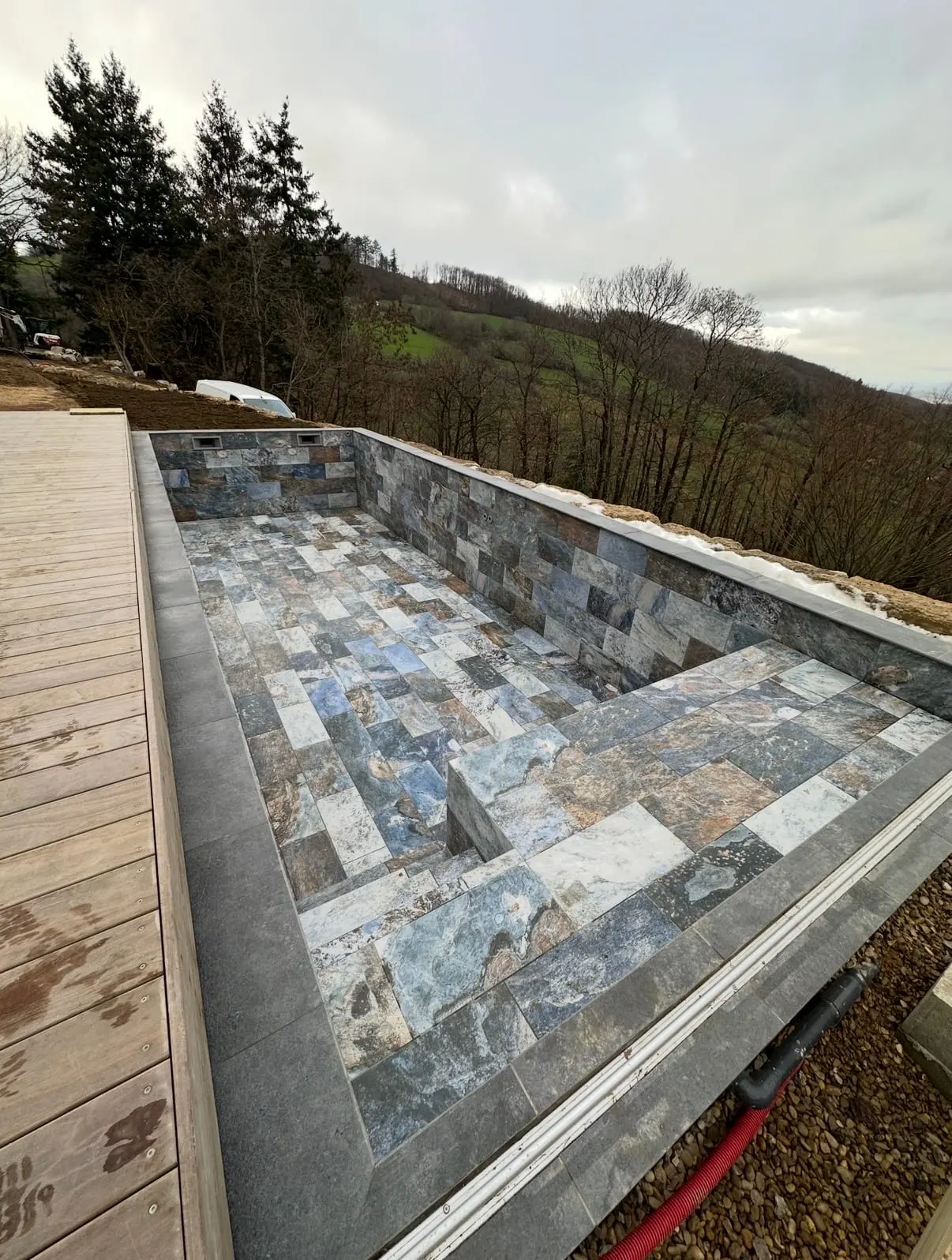 Piscine extérieure vide revêtue de carreaux en pierre aux nuances grises et bleutées, bordée d’une terrasse en bois face à un paysage boisé