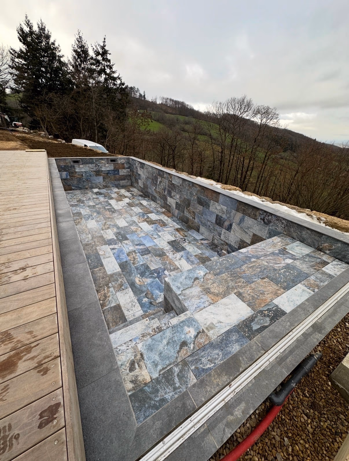 Piscine extérieure vide revêtue de carreaux en pierre aux nuances grises et bleutées, bordée d’une terrasse en bois face à un paysage boisé