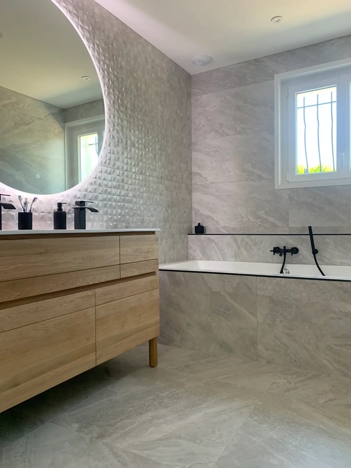Salle de bain avec carrelage gris effet pierre, mur texturé, baignoire et meuble vasque en bois clair